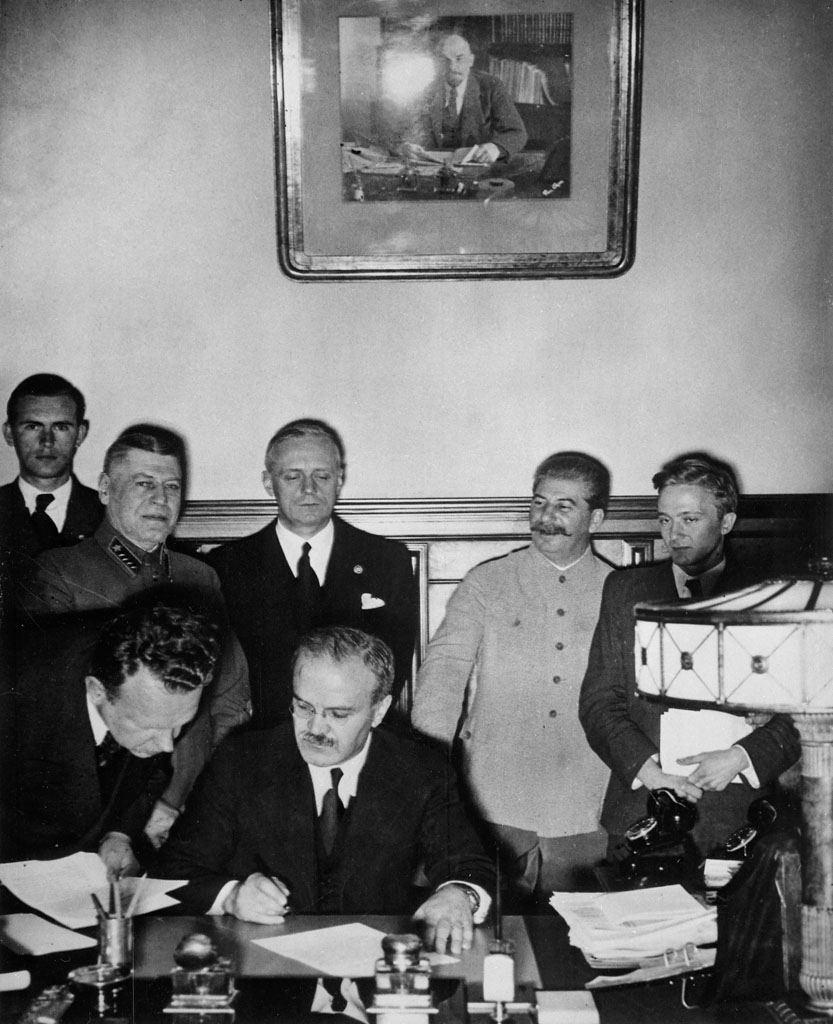 Molotov-Ribbentrop Pact, Vyacheslav Molotov, Russian foreign minister, signs the non-aggression pact negotiated between Soviet Russia and Germany, at the Kremlin, Moscow. Standing behind him is his German counterpart Joachim von Ribbentrop (left), and Joseph Stalin (centre). Image: public domain